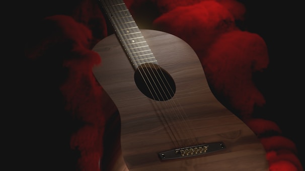 An acoustic guitar with a wooden finish is surrounded by dark shadows interspersed with vivid red smoke or fabric in the background. The lighting creates a dramatic contrast, highlighting the guitar's strings and body.