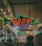 Neon signs indicating 'Market Grill' and 'Athenian Lunch Seafood' are visible, hanging from the ceiling in a bustling indoor market corridor. The signs are colorful, shaping a fish and arrow, respectively. Below, people are milling around, suggesting a busy, vibrant atmosphere. The lighting is warm and slightly dim.
