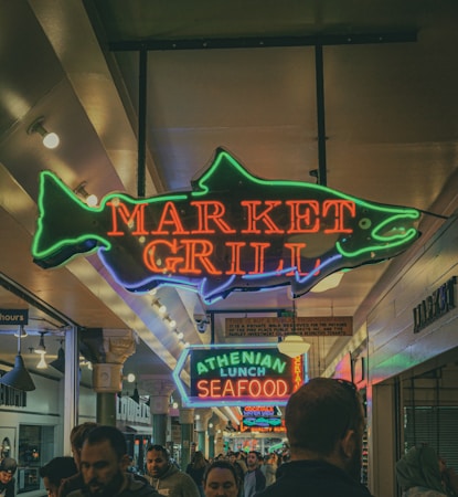 Neon signs indicating 'Market Grill' and 'Athenian Lunch Seafood' are visible, hanging from the ceiling in a bustling indoor market corridor. The signs are colorful, shaping a fish and arrow, respectively. Below, people are milling around, suggesting a busy, vibrant atmosphere. The lighting is warm and slightly dim.