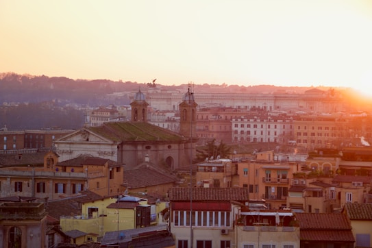 A picturesque view of a historic city at sunset, with an array of old buildings topped with terracotta rooftops. In the background, a prominent church with domes is visible, surrounded by other classic architecture under a warm, golden sky.