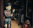 A wooden or ceramic sculpture of a person appears intricately detailed, painted in earthy tones and positioned next to a smaller bust. The scene seems to be inside a shop or gallery with various other sculptures and artifacts visible in the dimly-lit background.