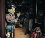 A wooden or ceramic sculpture of a person appears intricately detailed, painted in earthy tones and positioned next to a smaller bust. The scene seems to be inside a shop or gallery with various other sculptures and artifacts visible in the dimly-lit background.