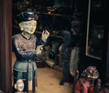 A wooden or ceramic sculpture of a person appears intricately detailed, painted in earthy tones and positioned next to a smaller bust. The scene seems to be inside a shop or gallery with various other sculptures and artifacts visible in the dimly-lit background.