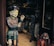 A wooden or ceramic sculpture of a person appears intricately detailed, painted in earthy tones and positioned next to a smaller bust. The scene seems to be inside a shop or gallery with various other sculptures and artifacts visible in the dimly-lit background.