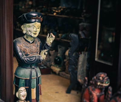 A wooden or ceramic sculpture of a person appears intricately detailed, painted in earthy tones and positioned next to a smaller bust. The scene seems to be inside a shop or gallery with various other sculptures and artifacts visible in the dimly-lit background.