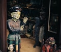 A wooden or ceramic sculpture of a person appears intricately detailed, painted in earthy tones and positioned next to a smaller bust. The scene seems to be inside a shop or gallery with various other sculptures and artifacts visible in the dimly-lit background.