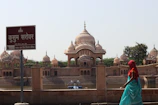 A serene view of Kusum Sarovar featuring ornate domed structures and their reflections in the water. A sign with Hindi text stands in the foreground, and a person in colorful traditional attire is seen walking along the water's edge. The ambiance is peaceful with a clear sky overhead.