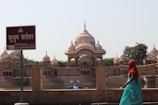 A serene view of Kusum Sarovar featuring ornate domed structures and their reflections in the water. A sign with Hindi text stands in the foreground, and a person in colorful traditional attire is seen walking along the water's edge. The ambiance is peaceful with a clear sky overhead.