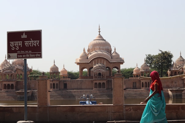 A serene view of Kusum Sarovar featuring ornate domed structures and their reflections in the water. A sign with Hindi text stands in the foreground, and a person in colorful traditional attire is seen walking along the water's edge. The ambiance is peaceful with a clear sky overhead.