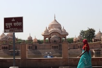 A serene view of Kusum Sarovar featuring ornate domed structures and their reflections in the water. A sign with Hindi text stands in the foreground, and a person in colorful traditional attire is seen walking along the water's edge. The ambiance is peaceful with a clear sky overhead.