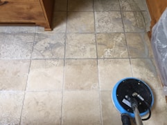 A tiled floor with visible dirt and cleaning solution being cleaned by a circular floor cleaning machine. The tiles are light brown with noticeable dirt in the grout lines. The cleaning machine has a blue and black circular head connected to a metal handle and hose.