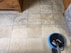 A tiled floor with visible dirt and cleaning solution being cleaned by a circular floor cleaning machine. The tiles are light brown with noticeable dirt in the grout lines. The cleaning machine has a blue and black circular head connected to a metal handle and hose.
