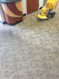 A patterned carpet covers most of the floor, with a yellow floor cleaning machine partially visible on the right. The pattern on the carpet appears to be a mix of geometric and abstract shapes. A section of a wooden counter with a curved design is on the left side.