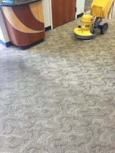 A patterned carpet covers most of the floor, with a yellow floor cleaning machine partially visible on the right. The pattern on the carpet appears to be a mix of geometric and abstract shapes. A section of a wooden counter with a curved design is on the left side.