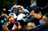 A large crowd of people holding and using cameras and smartphones, eagerly taking photographs of something off-frame. The scene is bustling and lively, with individuals of different ages intently focused on capturing the event. A man in the foreground is using a professional camera, demonstrating the serious interest in photography among the group.