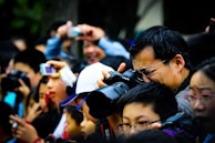 A large crowd of people holding and using cameras and smartphones, eagerly taking photographs of something off-frame. The scene is bustling and lively, with individuals of different ages intently focused on capturing the event. A man in the foreground is using a professional camera, demonstrating the serious interest in photography among the group.