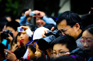 A large crowd of people holding and using cameras and smartphones, eagerly taking photographs of something off-frame. The scene is bustling and lively, with individuals of different ages intently focused on capturing the event. A man in the foreground is using a professional camera, demonstrating the serious interest in photography among the group.
