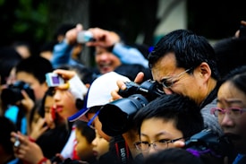 A large crowd of people holding and using cameras and smartphones, eagerly taking photographs of something off-frame. The scene is bustling and lively, with individuals of different ages intently focused on capturing the event. A man in the foreground is using a professional camera, demonstrating the serious interest in photography among the group.