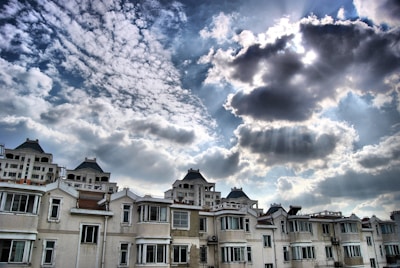 A row of residential buildings with multiple windows and balconies stands under a partly cloudy sky. Sunlight peeks through the clouds, casting rays across the scene. The architecture features white facades and dark, sloped roofs.
