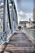 A metal bridge with intricate lattice structures spans across a wide pathway made of wooden planks, leading towards a bustling city skyline. A group of people walks along the bridge, with some buildings visible in the background under a partly cloudy sky.