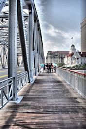 A metal bridge with intricate lattice structures spans across a wide pathway made of wooden planks, leading towards a bustling city skyline. A group of people walks along the bridge, with some buildings visible in the background under a partly cloudy sky.