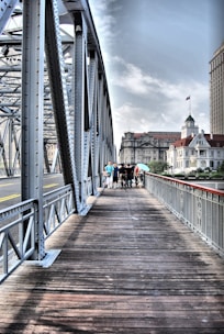 A metal bridge with intricate lattice structures spans across a wide pathway made of wooden planks, leading towards a bustling city skyline. A group of people walks along the bridge, with some buildings visible in the background under a partly cloudy sky.