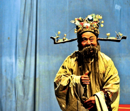 A person dressed in traditional Chinese opera attire, featuring an ornate headdress adorned with jewels and embellishments. The costume includes an elaborate robe with intricate patterns, complemented by a long, flowing beard. The background is a plain blue curtain, highlighting the vibrant colors of the attire.