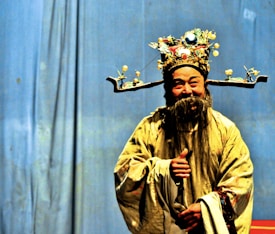 A person dressed in traditional Chinese opera attire, featuring an ornate headdress adorned with jewels and embellishments. The costume includes an elaborate robe with intricate patterns, complemented by a long, flowing beard. The background is a plain blue curtain, highlighting the vibrant colors of the attire.