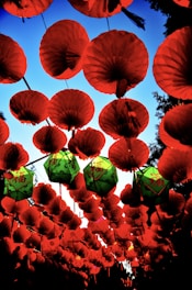 A vibrant arrangement of red lanterns hanging against a clear blue sky. Several green lanterns with Chinese characters are interspersed among the red ones, creating a striking contrast. The lanterns are suspended in rows, forming a canopy that evokes a festive and cultural ambiance.