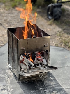 A small, stainless steel portable stove is set up outdoors with a fire burning inside. The flames are vibrant and dancing above the charred wood and ashes, providing warmth and cooking potential. The setting is natural, with a grassy ground and some blurred background elements suggesting a campsite.
