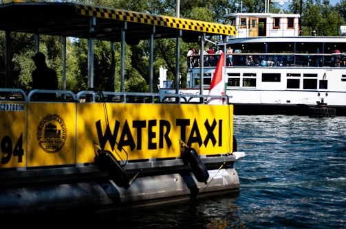 A bright yellow water taxi is docked on a body of water, with the words 'WATER TAXI' prominently displayed on its side. In the background, a larger ferry is filled with people, indicating a busy day on the water. The scene is framed by lush green trees, suggesting a natural and vibrant setting.