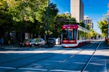 A modern red and white streetcar is traveling along tracks lined with trees in an urban setting. Cars are parked along the street, and buildings rise in the background. The day is bright and clear, casting defined shadows on the pavement.