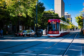 A modern red and white streetcar is traveling along tracks lined with trees in an urban setting. Cars are parked along the street, and buildings rise in the background. The day is bright and clear, casting defined shadows on the pavement.