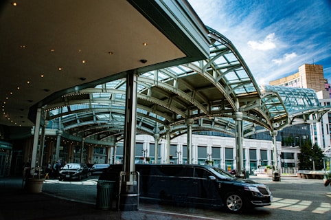A modern architectural structure with a glass and metal canopy covering an outdoor area. Several black vehicles, including a limousine, are parked beneath the canopy. The buildings in the background have a contemporary design with large windows. The scene is brightly lit with clear blue skies overhead.