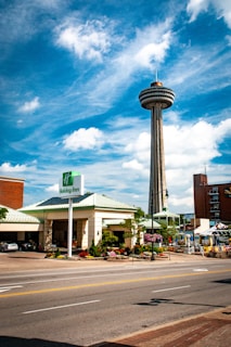 A tall observation tower with a circular platform at the top rises against a backdrop of a bright blue sky with scattered white clouds. Below, a Holiday Inn hotel with a green roof is visible, along with surrounding streets and sidewalks, featuring colorful flowers and greenery. Multiple cars are parked in the shaded area outside the hotel.