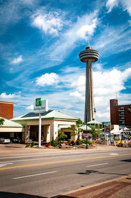 A tall observation tower with a circular platform at the top rises against a backdrop of a bright blue sky with scattered white clouds. Below, a Holiday Inn hotel with a green roof is visible, along with surrounding streets and sidewalks, featuring colorful flowers and greenery. Multiple cars are parked in the shaded area outside the hotel.