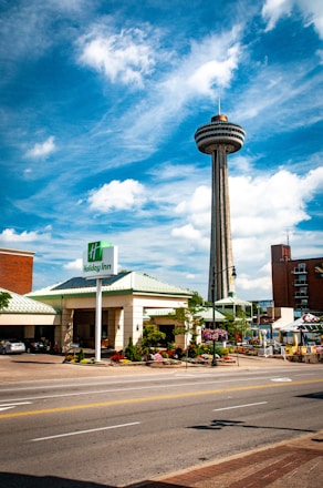 A tall observation tower with a circular platform at the top rises against a backdrop of a bright blue sky with scattered white clouds. Below, a Holiday Inn hotel with a green roof is visible, along with surrounding streets and sidewalks, featuring colorful flowers and greenery. Multiple cars are parked in the shaded area outside the hotel.