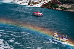 Two boats navigate on a river near a waterfall, with a prominent rainbow arcing over the water. One boat is closer to the viewer and is filled with people wearing blue ponchos, while the other, further away, has passengers wearing red. The background shows rocky cliffs and cascading water with lush greenery on the shoreline.