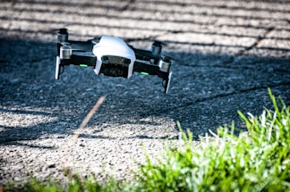 A small drone with a white body and black propellers flies above a textured, sunlit concrete surface, casting shadows on the ground. Green grass is visible in the foreground, adding contrast to the industrial feel of the setting.