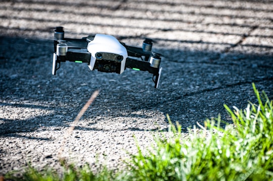 A small drone with a white body and black propellers flies above a textured, sunlit concrete surface, casting shadows on the ground. Green grass is visible in the foreground, adding contrast to the industrial feel of the setting.