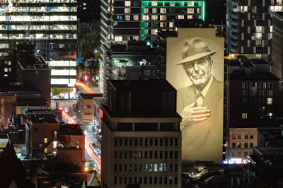 A large mural of an elderly man wearing a suit and hat is prominently displayed on the side of a building in an urban area. The man is depicted with his hand on his chest. The cityscape is illuminated by artificial lights, with various buildings and streets visible in the background.