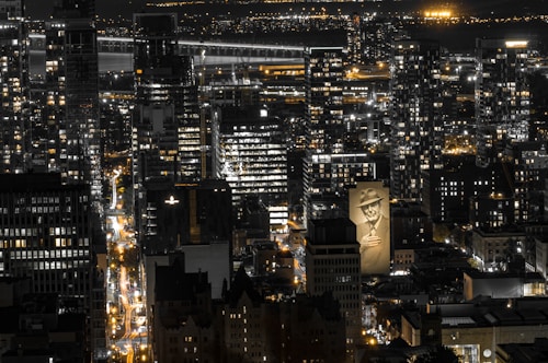 An urban nighttime scene with a densely packed cityscape featuring numerous illuminated skyscrapers and buildings. Amidst the city lights, a large mural of a man wearing a hat and holding a cigarette is prominently visible on one of the buildings.