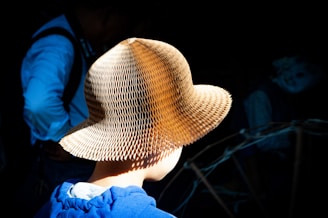 A person wearing a textured straw hat stands in focus against a dark background. The hat is intricately designed with a honeycomb pattern that catches the light, creating a striking visual contrast. The person wears a blue garment, partially visible, adding color to the otherwise shadowy setting.