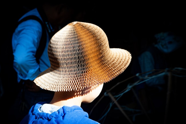 A person wearing a textured straw hat stands in focus against a dark background. The hat is intricately designed with a honeycomb pattern that catches the light, creating a striking visual contrast. The person wears a blue garment, partially visible, adding color to the otherwise shadowy setting.