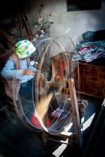 A person is seated at a spinning wheel, actively working with the machinery. The surroundings include a rustic wooden interior with cotton plants and neatly folded textiles. The motion of the spinning creates a blurred effect, conveying action and focus.