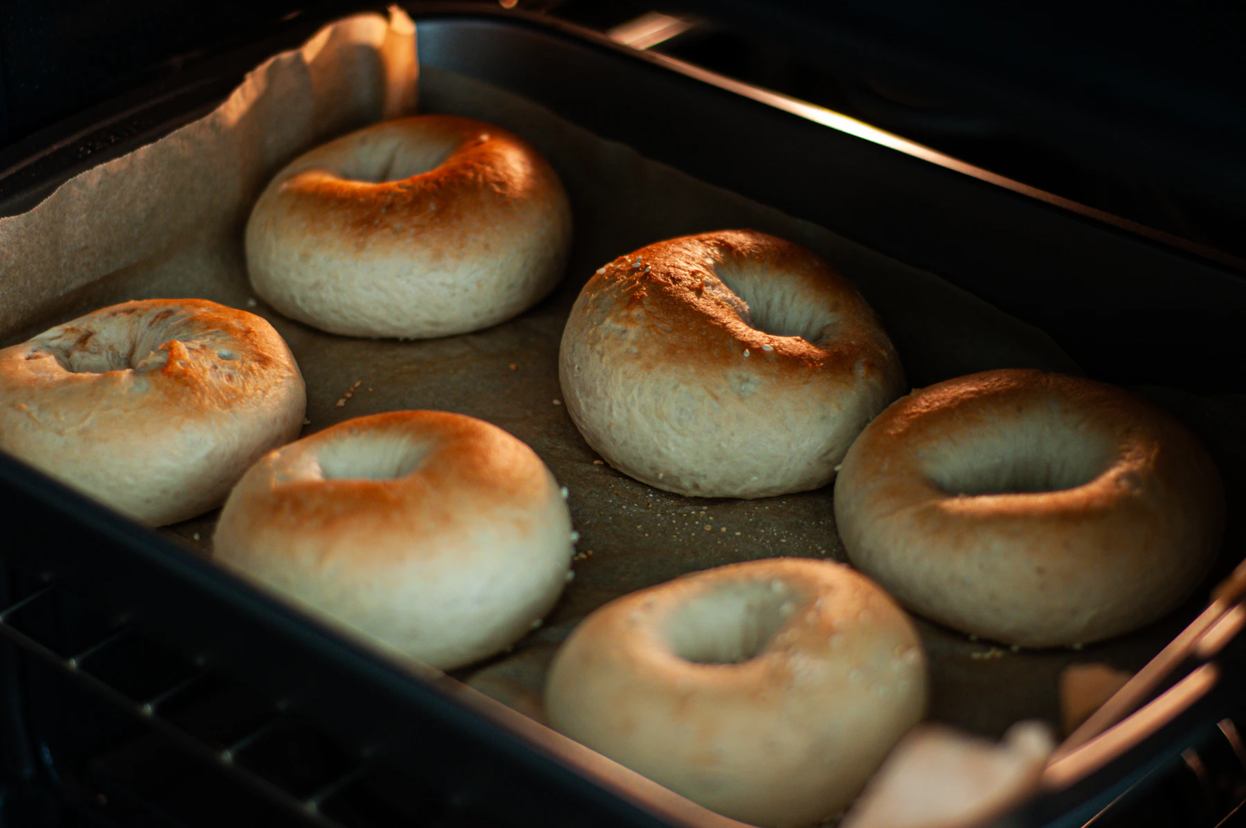 Pale bagels browning on a parchment-lined tray inside a lit oven