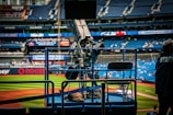 A professional-grade video camera is mounted on a platform overlooking a baseball field. The stadium seats in the background are filled with some spectators, and various advertisements are visible around the field. A staff member wearing a jacket with 'STAFF' written on the back is positioned near the camera.