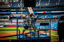 A professional-grade video camera is mounted on a platform overlooking a baseball field. The stadium seats in the background are filled with some spectators, and various advertisements are visible around the field. A staff member wearing a jacket with 'STAFF' written on the back is positioned near the camera.