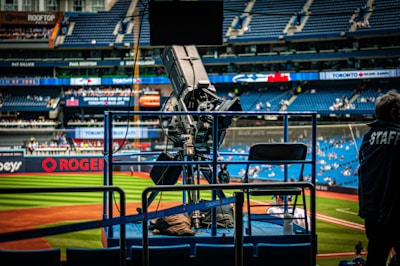 A professional-grade video camera is mounted on a platform overlooking a baseball field. The stadium seats in the background are filled with some spectators, and various advertisements are visible around the field. A staff member wearing a jacket with 'STAFF' written on the back is positioned near the camera.