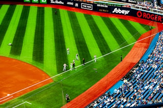 A baseball field with players in uniform stands ready near the sideline on the lush green grass. The stadium is partially filled with spectators in the seating area, and various advertisements are displayed on the perimeter fence.